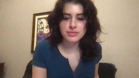 Media: Video of a young woman with long, curly black hair and light skin, wearing a navy blue V-neck shirt. Background includes a beige wall with a framed colorful painting.