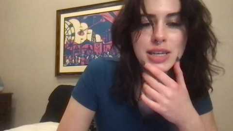 Media: Video of a young woman with wavy black hair, wearing a blue shirt, holding her chin, in a bedroom with a colorful abstract painting and a dark wooden nightstand.