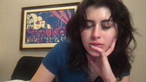 Media: A video of a young woman with shoulder-length, wavy black hair, resting her chin on her hand, wearing a dark blue t-shirt. She is looking down, slightly smiling. Background includes a framed, colorful abstract painting and beige wall.