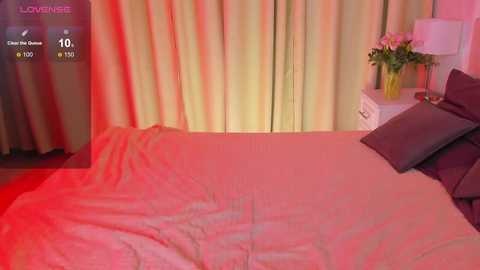 Media: A video of a bedroom with a beige bed, two pillows, a white nightstand, and a vase of flowers under soft, pink lighting.