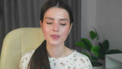 Media: Video of a young woman with fair skin, dark hair, wearing a floral shirt, seated indoors with green potted plants and grey curtains in the background.