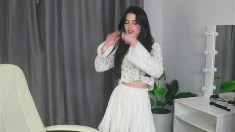 Media: Video of a young woman with long black hair, wearing a white lace crop top and pleated skirt, dancing joyfully in a modern, minimalist room with gray curtains, white furniture, and a green plant.