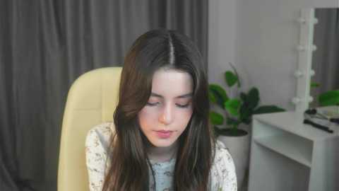 Media: Video of a young woman with long, dark brown hair, fair skin, and closed eyes, wearing a white floral-patterned top. Background features a light-colored chair, grey curtains, a white vanity table with green plants and makeup items.