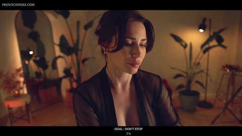 Media: A video of a fair-skinned woman with dark hair, wearing a black sheer top, in a dimly lit, warmly colored room with potted plants and arched doorways.