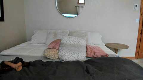 Media: Video of a minimalist, cozy bedroom with a white bed, pink and white pillows, a round mirror, and a wooden nightstand. A person with a black hoodie is partially visible at the bottom, lying on a gray blanket.