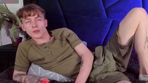 Media: Video of a young, fair-skinned, red-haired man with tattoos, wearing olive-green shirt and shorts, lounging on a dark couch, with a potted plant in the background.