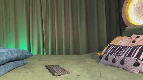 Media: A video of a cozy bedroom with green curtains, a yellow light, a blue pillow, a striped pillow, and a keyboard on a green bedspread.