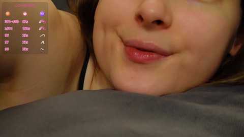 Media: A close-up video of a smiling Asian woman with light skin, lying on a gray pillow, showing her lips and nose. The background features a digital display with icons and text.