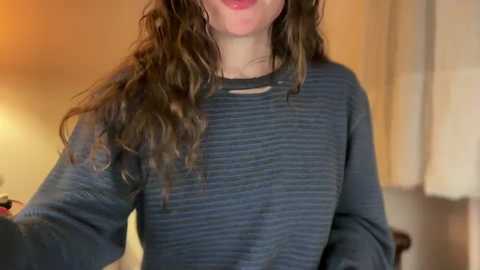 Media: Video of a woman with long, wavy brown hair, wearing a dark grey, striped, long-sleeved sweater, smiling. Background features a blurred indoor setting with soft lighting and beige walls.