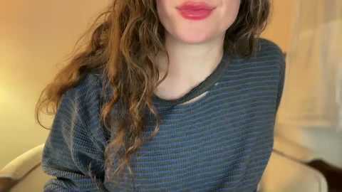 Media: A video of a young woman with long, curly brown hair and fair skin, wearing a dark blue, striped sweater, partially visible, against a blurred, warm-toned background.