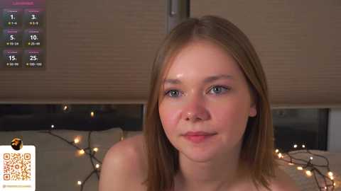 Media: Video of a young, fair-skinned woman with straight, light brown hair and blue eyes, smiling slightly. She has a smooth complexion. Background features a beige wall with a digital clock displaying time and temperature, and a string of warm, yellow fairy lights.