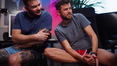 Media: Video of two tattooed, muscular men in casual clothes, one in blue and the other in gray, sitting on a black chair, chatting indoors.