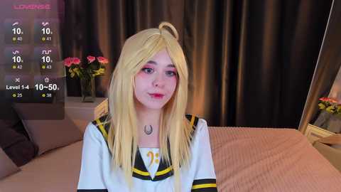 Media: Video of a young woman with long blonde hair, wearing a white sailor uniform with black and yellow accents, sitting on a bed with pink sheets. Background includes a mirror and floral arrangement.