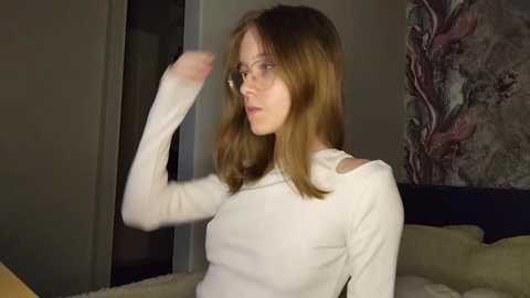 Media: Video of a young woman with shoulder-length, straight, light brown hair, wearing a white long-sleeve top, adjusting her hair in a dimly lit room with a floral tapestry on the wall.