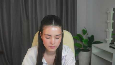 Media: Video of a young woman with fair skin, long dark hair, and closed eyes, sitting on a beige chair in a room with gray curtains, a white shelf, and a green plant.