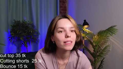 Media: Video of a fair-skinned woman with shoulder-length brown hair, wearing a purple top, standing indoors with a fern plant, green curtains, and a lamp in the background. Text overlays indicate she is cutting shorts, 25.5 k, and bounce 15.