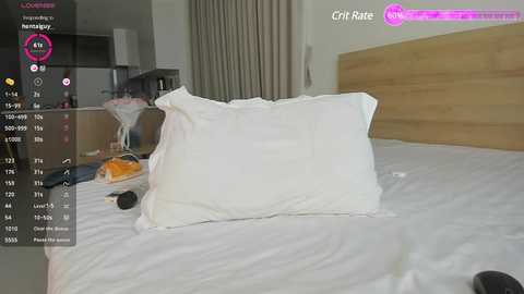 Media: Video of a messy bedroom with a white pillow on a bed, orange pillow nearby, and a wooden headboard. Background shows a kitchen counter with items and beige curtains.
