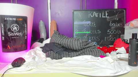 Media: Video of a messy bedroom with a pile of gray and black striped clothes on a bed, a black chalkboard with Karlie written on it, and a white SisWet bottle.