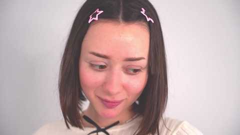 Media: Video of a young woman with straight, shoulder-length brown hair, styled with pink star hair clips. She has fair skin, light makeup, and is wearing a white top. She gazes downward with a soft smile.