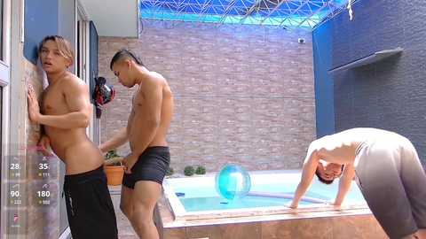 Video of two shirtless, muscular men in swim shorts standing inside a pool house, with a third man, also shirtless, bending over the pool.