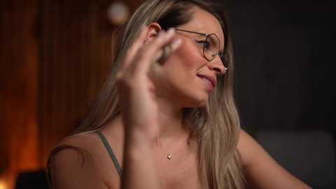 Media: A video of a blonde woman with glasses and a green top, adjusting her hair, in a dimly-lit room with blurred, warm-toned background.