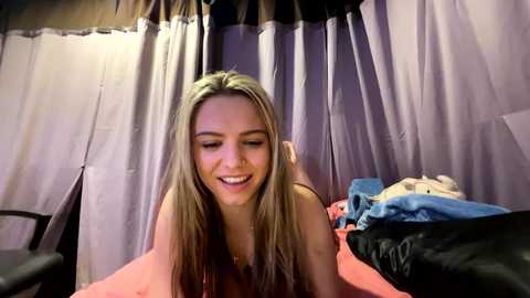 Media: Video of a smiling, slender, blonde woman with fair skin, wearing a sleeveless top, lying on a bed with a pink sheet. The background features white and purple curtains and disheveled clothes.