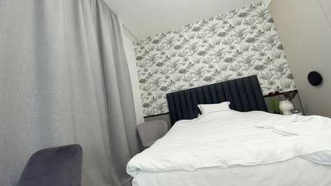 Media: Video of a modern bedroom with a tufted black headboard, white bedding, and gray curtains. The background features a monochrome floral wallpaper.