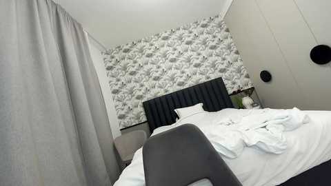 Media: Video of a minimalist bedroom with a white headboard, white bedding, gray curtains, and a white wall with floral pattern.