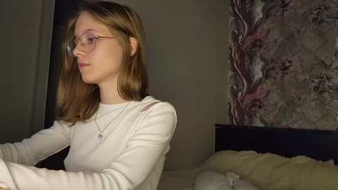 Media: Video of a young woman with light skin and straight brown hair, wearing glasses and a white long-sleeved shirt, sitting on a bed in a dimly lit bedroom with floral wallpaper.