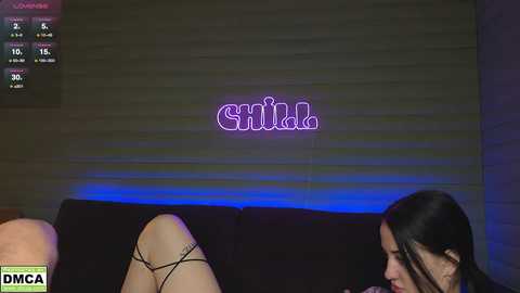 Media: Video of a woman with long black hair, wearing a black thong, sitting on a couch with a \"Smile\" neon sign behind her, in a dimly lit room.