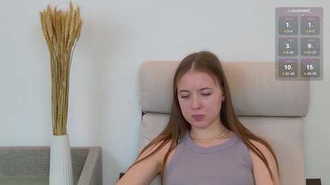 A video of a young woman with long brown hair, wearing a sleeveless lavender top, sitting on a beige chair in a minimalist room.