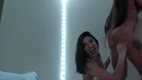 Media: A video of a smiling nude woman with long dark hair, holding the buttocks of another nude woman, against a dimly lit, white-walled background with glowing LED lights.