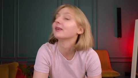 Media: A video of a Caucasian woman with fair skin and shoulder-length blonde hair, wearing a light pink t-shirt, smiling and tilting her head back. The background features teal walls, a modern orange couch, and a black wall-mounted shelf.