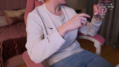 Media: Video of a fair-skinned woman with red hair, wearing a white Nike sweatshirt, blue jeans, and pink gaming chair. She holds a controller, and the background shows a pink bed and curtains.
