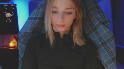 Media: Video of a blonde woman with fair skin, wearing a black jacket, lying on a blue plaid blanket in a dimly lit room with a fireplace and a candle.