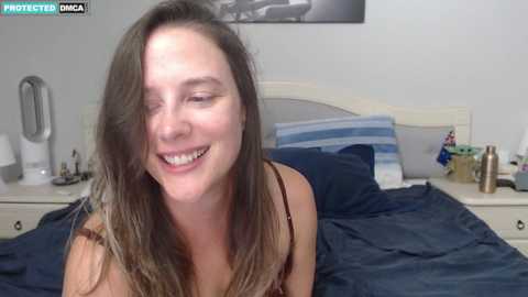 Media: A video of a young woman with long brown hair, wearing a brown top, smiling on a bed with blue sheets, in a bedroom with a light-colored headboard.