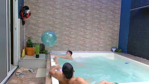 Video of a young man in a bathtub, with a blue bubble toy, surrounded by a tiled shower area, and a person outside the shower door watching.