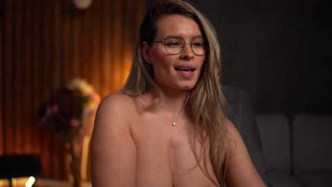 Media: Video of a topless woman with long blonde hair, wearing glasses, smiling, in a dimly lit room with wooden paneling and a vase of flowers.