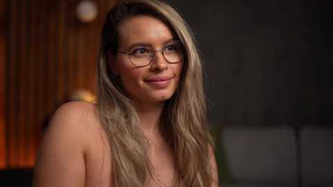 Media: Video of a smiling, topless woman with long, wavy blonde hair, wearing glasses, against a blurred wooden and dark wall background.