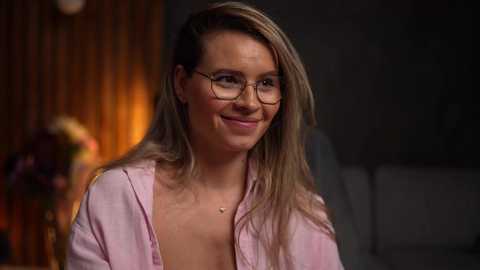 Media: Video of a smiling, light-skinned woman with straight, shoulder-length blonde hair, wearing glasses and a pink button-up shirt, indoors with dim lighting and blurred background.