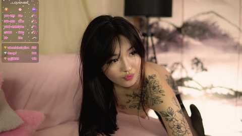 Media: Video of an East Asian woman with long black hair and tattoos, lying on a pink couch, wearing a black bra. Background includes a lamp and blurred furniture.