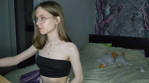 Media: Video of a young, slim, fair-skinned woman with straight, light brown hair, wearing glasses and a black strapless top, sitting on a bed with a white comforter, next to a stuffed duck, in a dimly lit room.