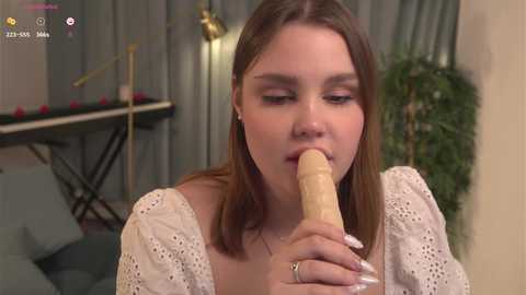 Media: Video of a young woman with fair skin, light brown hair, and light makeup, performing oral sex on a large, flesh-colored dildo in a dimly lit room with a pool table and green curtains in the background.