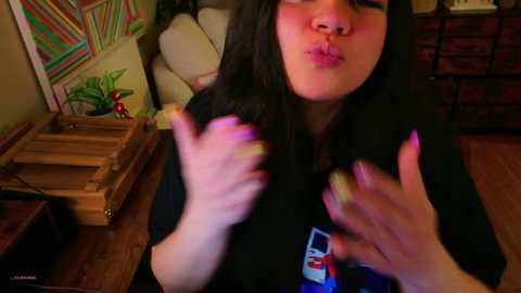 Media: Video of a young woman with long black hair, fair skin, and dark eyes, making a kissing face. She wears a black T-shirt and has painted nails. Background features colorful wall art, wooden shelves, and a brick fireplace.