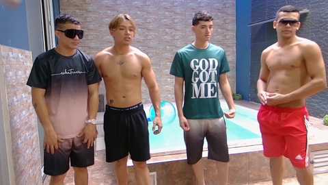 Video of four shirtless young men in swim trunks, standing near an indoor pool with blue water. One wears black shorts, another a green shirt with \"God of Me\" text, and the others red shorts.