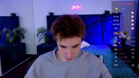 Media: Video of a young man with short brown hair, wearing a gray sweater, standing in a modern bedroom with a bed, plants, and a TV streaming a video game.