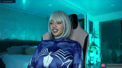 Media: Video of a woman with platinum blonde hair, wearing a Spider-Man-themed cape, seated in a gaming chair in a dimly lit room with blue lighting.