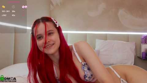 Media: Video of a young woman with long red hair, wearing a white floral bra and panties, lying on a bed with white pillows. The room has a modern, minimalist decor.