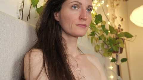 Media: Video of a fair-skinned woman with long brown hair, wearing a necklace, standing against a beige wall with green plants, and a soft-lit round lamp in the background.