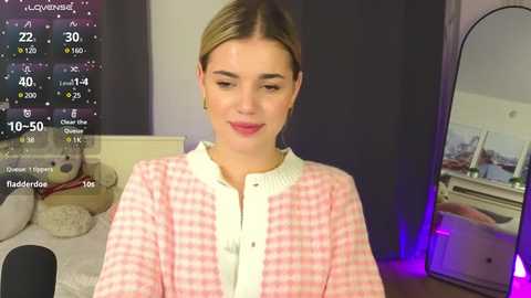 Media: Video of a light-skinned, blonde woman with a medium build in a pink gingham shirt, sitting in a cozy, dimly lit bedroom. Background includes a bed with plush toys, a mirror, and a digital clock displaying the time.
